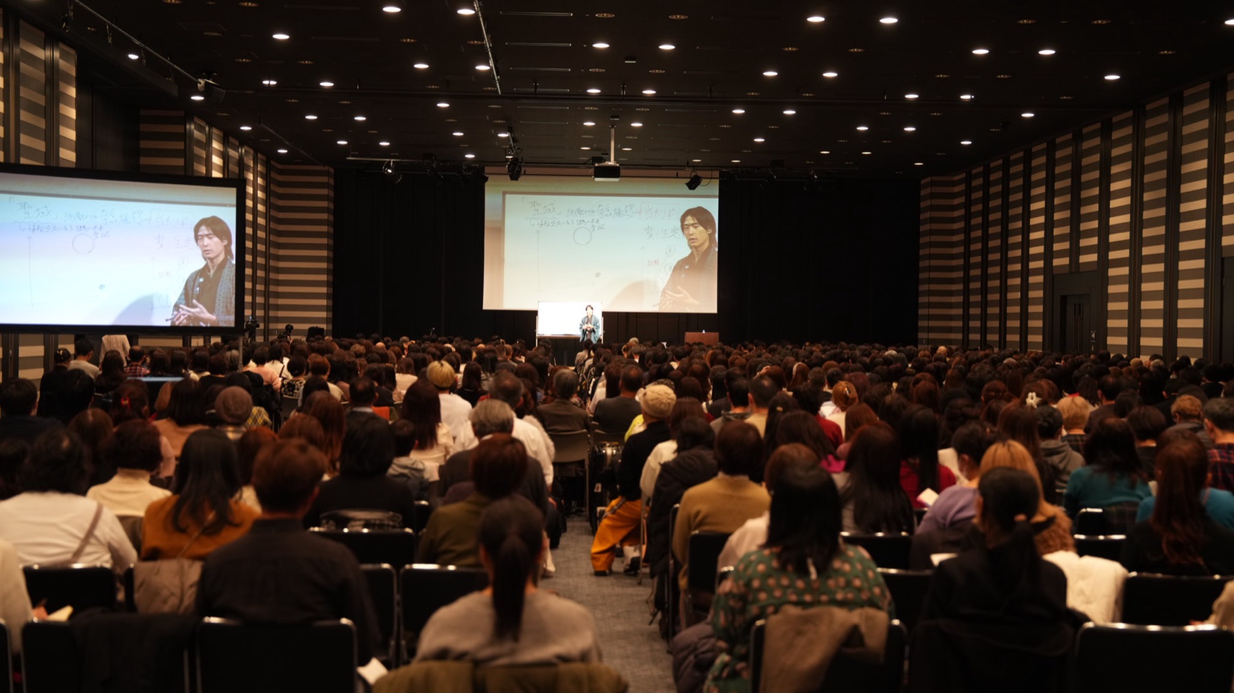 大型会場での講演の様子。満席の客席と登壇する代表